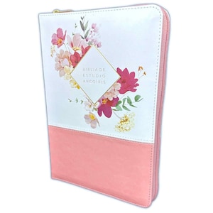 May include: A pink and white Bible cover with a floral design and the text "BIBLIA DE ESTUDIO ARCOIRIS". The cover has a zipper closure and a gold-colored zipper pull. The top half is white with a floral design, and the bottom half is solid pink.