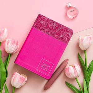 May include: A pink leather-bound journal with a floral design on the cover. The journal has the words "SANTA BIBLIA" printed on the front. The journal is surrounded by pink tulips and a small bottle of perfume.