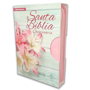 May include: A pink and white Bible with the text "Santa Biblia Quinceañera" on the cover. The cover features a floral design with pink lilies and a light blue wood-grain background. The Bible is encased in a clear plastic sleeve.