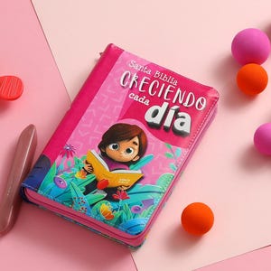 May include: A pink hardcover book with the title "Santa Biblia Creciendo cada dia" and an illustration of a girl reading a book in a garden.