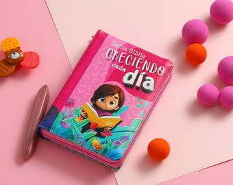 May include: A pink hardcover book with the title "Santa Biblia Creciendo cada dia" and an illustration of a girl reading a book in a garden.