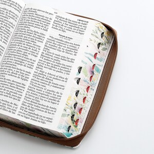 Puede incluir: Una Biblia de tapa dura de cuero marr&oacute;n con cierre de cremallera. La cubierta presenta un dise&ntilde;o floral con flores y hojas coloridas. Las p&aacute;ginas est&aacute;n abiertas en el libro de los Reyes, con texto en espa&ntilde;ol.