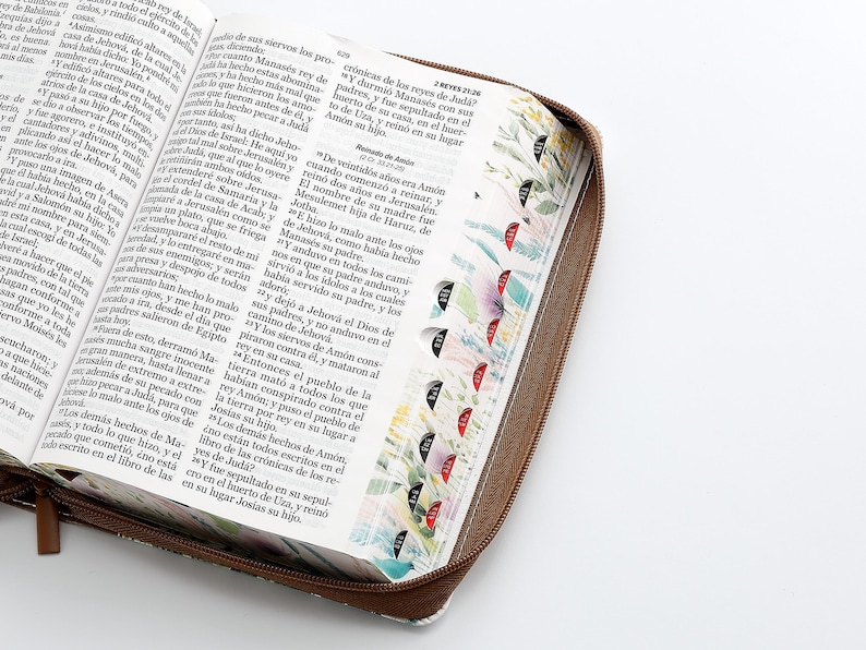 Puede incluir: Una Biblia de tapa dura de cuero marr&oacute;n con cierre de cremallera. La cubierta presenta un dise&ntilde;o floral con flores y hojas coloridas. Las p&aacute;ginas est&aacute;n abiertas en el libro de los Reyes, con texto en espa&ntilde;ol.
