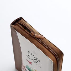 Puede incluir: Una funda de cremallera marr&oacute;n con un panel frontal blanco. El panel frontal tiene un dise&ntilde;o floral y el texto "Santa Biblia" en cursiva.