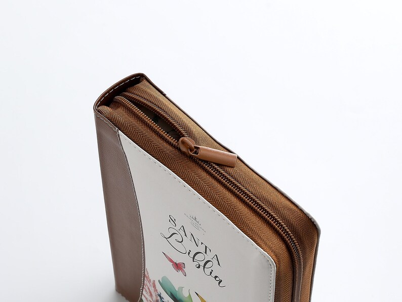 Puede incluir: Una funda de cremallera marr&oacute;n con un panel frontal blanco. El panel frontal tiene un dise&ntilde;o floral y el texto "Santa Biblia" en cursiva.