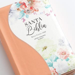 May include: A peach-colored leather-bound Bible with a floral design. The cover features a white background with pink, blue, and green flowers. The title "SANTA BIBLIA" is printed in black lettering, along with the text "Reina-Valera 1960" and "Letra Gigante - 14 puntos."