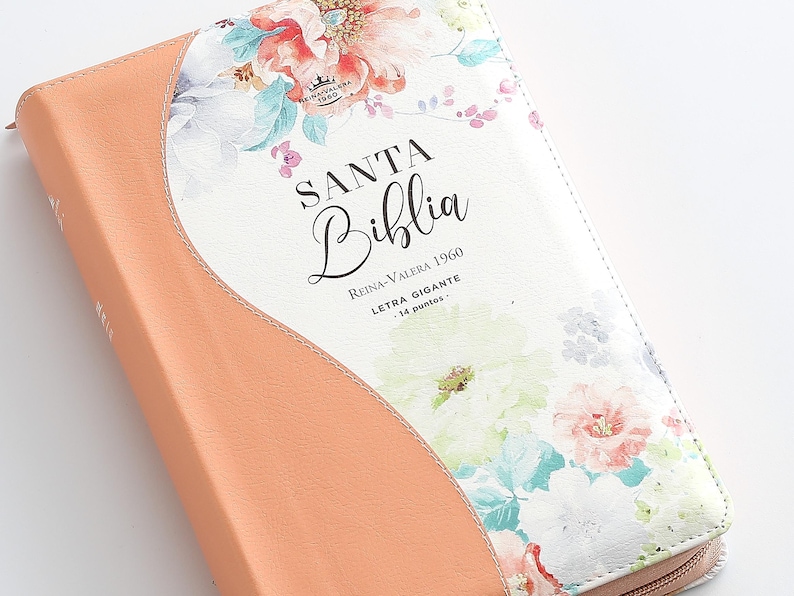 May include: A peach-colored leather-bound Bible with a floral design. The cover features a white background with pink, blue, and green flowers. The title "SANTA BIBLIA" is printed in black lettering, along with the text "Reina-Valera 1960" and "Letra Gigante - 14 puntos."