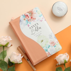 May include: A peach-colored leather-bound journal with a floral design on the cover. The cover reads "SANTA Biblia" with the text "Reina Valera 1960" and "Letra Gigante" underneath.