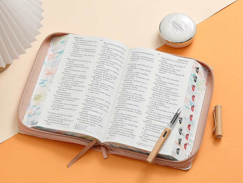 May include: A pink leather-bound journal with a zipper closure. The journal is open to reveal pages with Spanish text. A pen and a small round container are also visible.