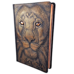 May include: A black Bible with a detailed embossed lion's face on the cover. The lion's mane and facial features are in shades of gold and brown. The text "SANTA BIBLIA" is embossed in gold.