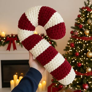 Puede incluir: Un gran cojín de bastón de caramelo hecho a mano con rayas rojas y blancas alternas. El cojín está hecho de un hilo grueso y texturizado. Se sostiene frente a un árbol de Navidad y una chimenea.
