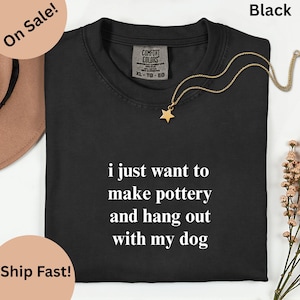 May include: Black t-shirt with the text "i just want to make pottery and hang out with my dog" in white. A gold necklace with a star pendant is draped on the shirt. The shirt is labeled XL-TG-EG.