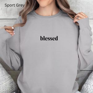 May include: Sport Grey sweatshirt with the word "blessed" printed in black. The sweatshirt is a light grey color and has a crew neck. The text is centered on the chest.