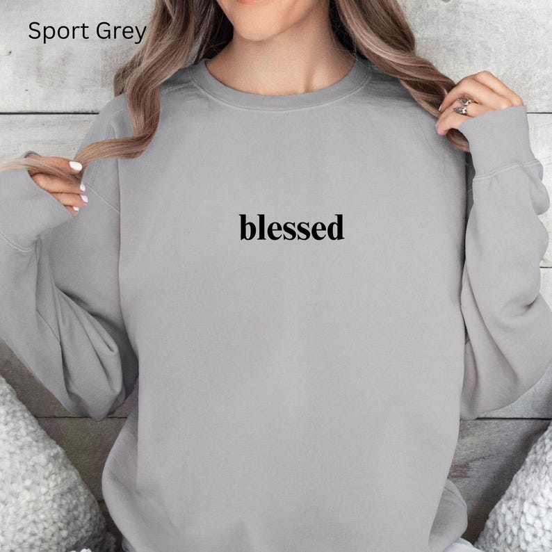 May include: Sport Grey sweatshirt with the word "blessed" printed in black. The sweatshirt is a light grey color and has a crew neck. The text is centered on the chest.