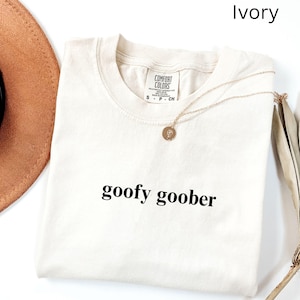 May include: Ivory-colored t-shirt with the words "goofy goober" printed in black. The shirt has a crew neck and short sleeves. A gold necklace with a pendant is draped on the shirt. A brown hat is partially visible.