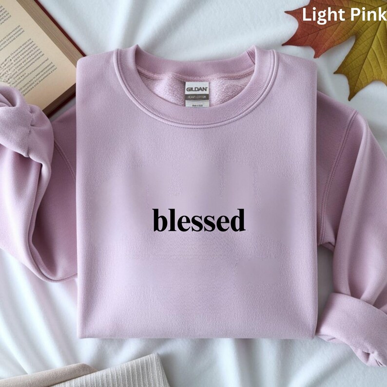May include: A light pink sweatshirt with the word "blessed" printed in black across the front. The sweatshirt has a crew neck and long sleeves. The brand tag is visible at the back of the neck. The sweatshirt is folded neatly.