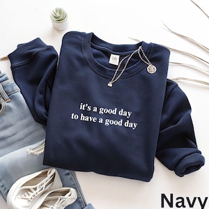 May include: Navy blue sweatshirt with the text "it's a good day to have a good day" in white. A silver necklace with a pendant rests on the sweatshirt. The word "Navy" is displayed in the bottom right corner. The sweatshirt is paired with blue jeans and white sneakers.
