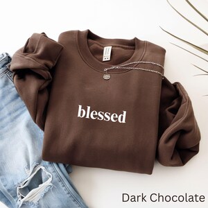 May include: A dark chocolate-colored sweatshirt with the word "blessed" in white lettering. A silver necklace with a lotus flower pendant rests on the sweatshirt. Light blue jeans are visible in the background.