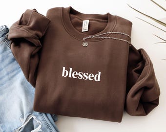 May include: A dark chocolate-colored sweatshirt with the word "blessed" in white lettering. A silver necklace with a lotus flower pendant rests on the sweatshirt. Light blue jeans are visible in the background.