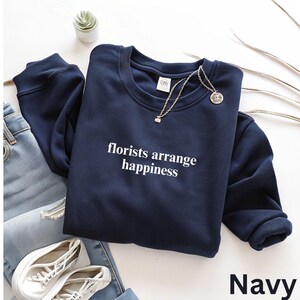 May include: Navy blue sweatshirt with the text "florists arrange happiness" in white. The crew neck sweatshirt is paired with light wash jeans and white sneakers. A silver necklace with a pendant is draped on the sweatshirt.