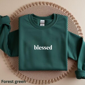 May include: Forest green sweatshirt with the word "blessed" in white block letters. The sweatshirt is displayed on a woven, circular placemat. The sleeves are cuffed at the wrists.