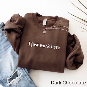 May include: A dark chocolate-colored sweatshirt with the phrase "i just work here" in white lettering. A silver necklace with a pendant is draped across the sweatshirt. Light blue jeans are visible on the left.