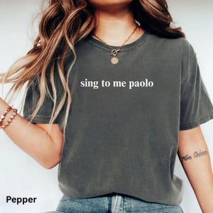 May include: A charcoal gray short-sleeve t-shirt featuring the white text "sing to me paolo" printed across the chest. The garment has a crew neckline and a relaxed fit, showcasing a solid, muted color tone throughout the fabric design.