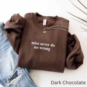 May include: A dark chocolate-colored sweatshirt with the text "miss never do no wrong" in white. A silver necklace with a lotus flower pendant is draped over the sweatshirt. Light blue jeans are visible in the background.