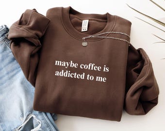 May include: A dark chocolate-colored sweatshirt with the text "maybe coffee is addicted to me" in white. A silver necklace with a small pendant is draped over the sweatshirt. Light blue jeans are visible on the left side of the image.