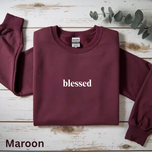 May include: Maroon crewneck sweatshirt with the word "blessed" in white script. The sweatshirt is folded, with the sleeves extending to the sides. The brand tag is visible at the back of the neck. The word "Maroon" is at the bottom.