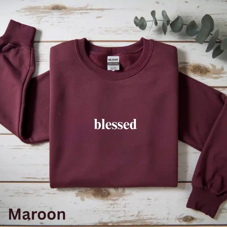 May include: Maroon crewneck sweatshirt with the word "blessed" in white script. The sweatshirt is folded, with the sleeves extending to the sides. The brand tag is visible at the back of the neck. The word "Maroon" is at the bottom.