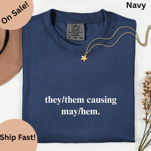 May include: Navy blue t-shirt with the text "they/them causing may/hem." A gold star pendant hangs on a gold chain. The shirt has a Comfort Colors tag. The words "On Sale!" and "Ship Fast!" are in the image.