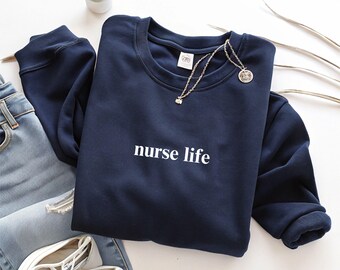 May include: Navy blue crewneck sweatshirt with the text "nurse life" in white. The sweatshirt is folded on a white surface next to a pair of light blue jeans, white sneakers, and a silver necklace. A small succulent plant is also visible.