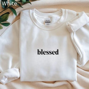 May include: White crewneck sweatshirt with the word "blessed" printed in black. The sweatshirt is made of a soft material and has a classic, casual style. The word "White" is in the top left corner.