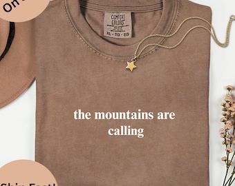 May include: Espresso-coloured t-shirt with the text "the mountains are calling." A gold-coloured star pendant hangs from a delicate chain. The shirt is folded and has a Comfort Colors tag. A sprig of flowers is on the right side of the shirt.