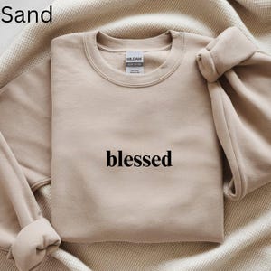 May include: Sand-colored sweatshirt with the word "blessed" printed in black. The sweatshirt is made of a soft material and has a crew neck. The sleeves are folded over. The sweatshirt is laying on a beige blanket.