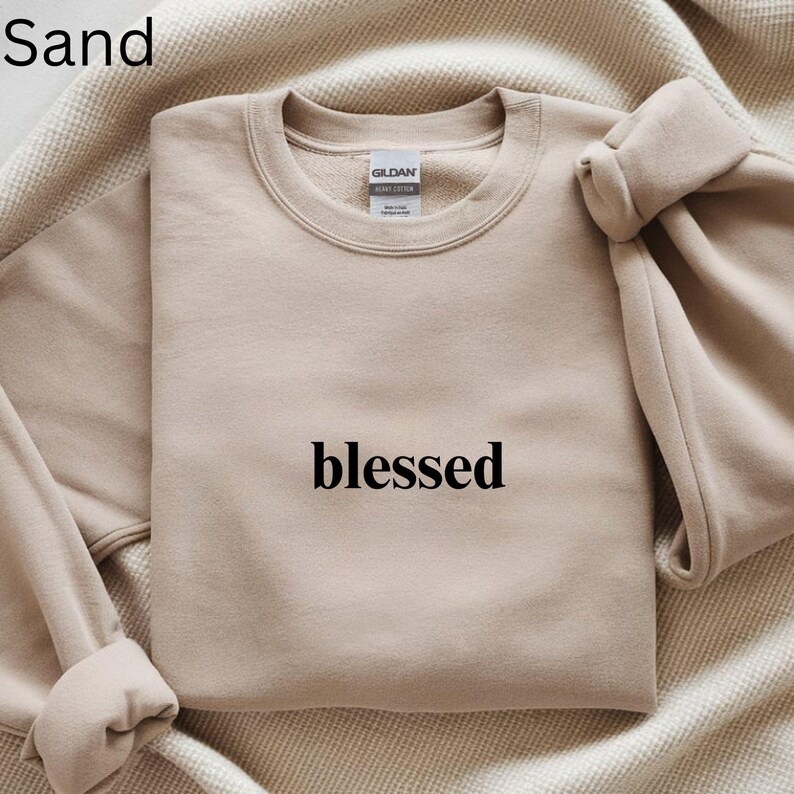 May include: Sand-colored sweatshirt with the word "blessed" printed in black. The sweatshirt is made of a soft material and has a crew neck. The sleeves are folded over. The sweatshirt is laying on a beige blanket.