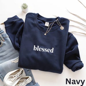 May include: A navy blue sweatshirt with the word "blessed" printed in white. A silver necklace with a small pendant rests on the sweatshirt. The word "Navy" is printed in white at the bottom right corner of the image. A pair of blue jeans and white sneakers are also visible.