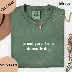 May include: A moss green t-shirt with the text "proud parent of a dramatic dog" in white. The shirt has a crew neck and a small tag on the upper left. A gold star pendant necklace is draped across the shirt.
