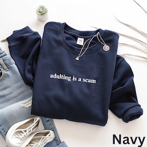 May include: Navy blue sweatshirt with the phrase "adulting is a scam" in white lettering. The sweatshirt is paired with light blue jeans, white sneakers, and a silver necklace. A small plant and decorative branches are also visible.