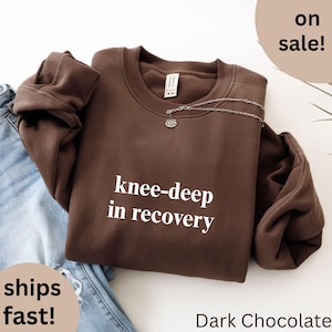 May include: Dark chocolate-colored sweatshirt with the text "knee-deep in recovery" in white. A silver necklace with a pendant is draped across the top. The sweatshirt is folded over a pair of blue jeans.