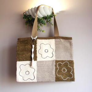 Puede incluir: Un bolso tote de patchwork con un diseño floral. El bolso presenta cuadrados de tela verde oliva, blanca y gris, cada uno con una flor bordada en blanco. Tiene asas marrones y un charm de borla blanca.