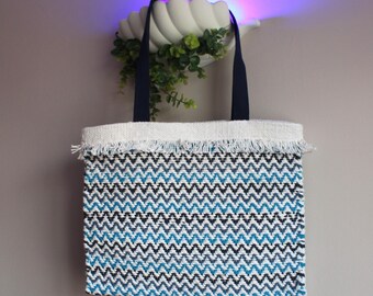 May include: Woven tote bag with a chevron pattern in blue, black, and white. The top edge has a white fringe, and the handles are navy blue. The bag is hanging on a wall.