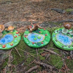 May include: Three miniature fairy garden scenes with ponds, mushrooms, and a tree stump. Each scene is set on a round, green base with red flowers and brown rocks.