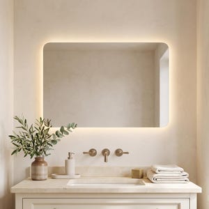 May include: A rectangular bathroom vanity with a cream-colored cabinet, a stone countertop, and an integrated sink. Above the sink is a wall-mounted mirror with rounded corners and integrated LED backlighting, paired with a bronze-toned faucet.