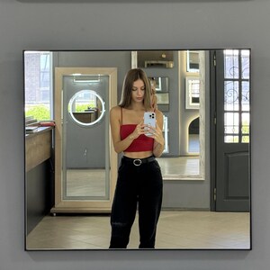 May include: A woman in a red crop top and black pants stands in front of a mirror, taking a selfie with a smartphone. The mirror is surrounded by a black frame and reflects the room behind her.