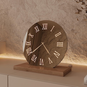 May include: A circular, reflective bronze-toned clock face featuring gold-colored Roman numerals and two thin, matching hands. The clock is mounted on a rectangular, light-brown wooden base, creating a stable, upright display for the timepiece.