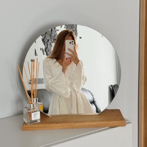 Può includere: Uno specchio rotondo con una base in legno. Lo specchio riflette una persona che indossa un vestito bianco e tiene uno smartphone.