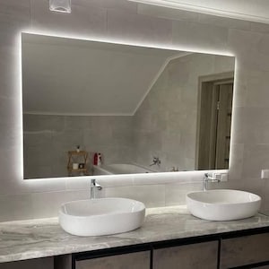 May include: A bathroom vanity with two white porcelain sinks and a large rectangular mirror with an illuminated border. The vanity has a dark grey countertop and three drawers with black metal frames.