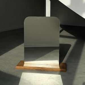 Puede incluir: Un espejo rectangular con esquinas superiores redondeadas está montado sobre una base de madera rectangular. El espejo refleja el entorno. La base de madera tiene un acabado de veta natural. El espejo es un objeto decorativo.